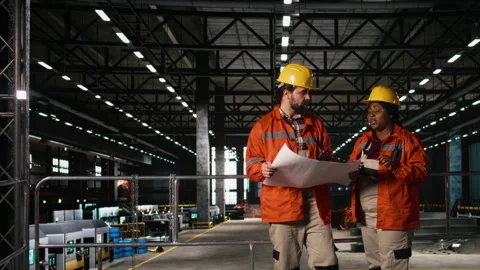 Diverse team of engineers studying blueprints and layout plans in machinery hall Stock Footage 330377974