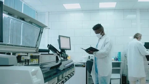 A diverse team watches robots work in laboratory, wearing masks and gowns 스톡 동영상 166665040
