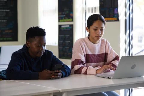 Diverse teens sitting in classroom at table in hoodie and pink sweater, using Stock Photos