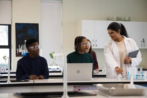 Diverse teens watching Indian instructor in lab coat pointing at test tubes with Stock Photos
