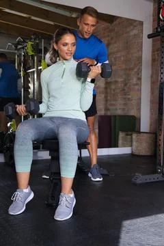 Diverse trainer spotting while client performing dumbbell curls wearing mint top Stock Photos