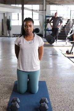 Diverse trainer watching client kneeling on blue mat with hex dumbbells near Stock Photos