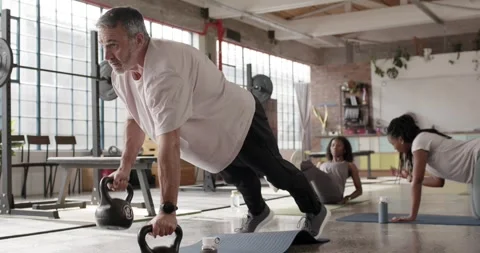 Diverse trio warming up in loft, man using kettlebells doing plank pushes then Stock Footage 337286780
