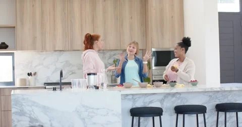 Diverse women chatting while prepping fruit on marble island in kitchen, tossing Stock Footage 337238744