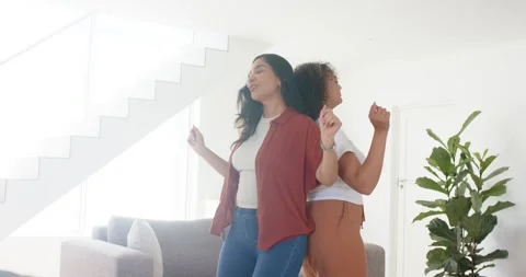 Diverse women friends smiling, syncing back-to-back moving at home by sofa rust 動画素材 340613490