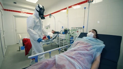 Doctor in a hazmat suit is checking temperature of a lying patient. Covid-19 Stock Footage 128084933