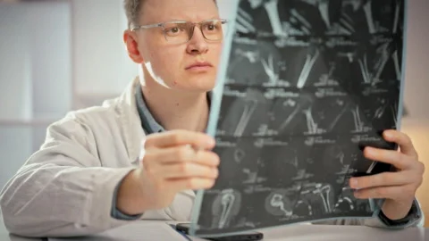 A doctor leans over a table in a bright medical office scrutinizing MRI scan Stock Footage 345526885