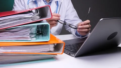 Doctor sitting at desk. Reviewing a stack of ring binders filled with documents Stock Footage 324768502