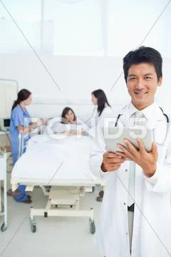 Doctor Smiling While Holding A Tablet As His Colleagues Treat A Patient