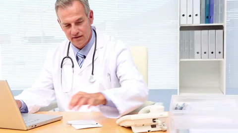 Doctor talking on phone and using laptop at his desk 動画素材 34077145