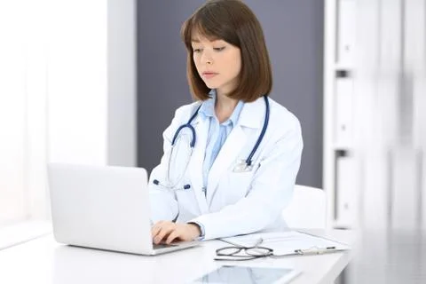 Doctor typing on laptop computer while sitting at the white table in hospital Stockfoto's