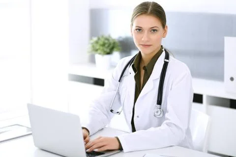 Doctor typing on laptop computer while sitting at the desk in hospital office Stock Photos