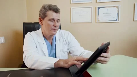 A doctor uses a tablet while sitting at a desk in his office. Stock Footage 256157314