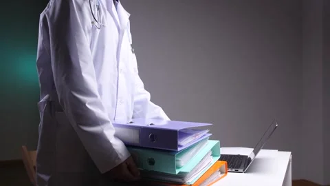 Doctor in a white coat carrying a stack of files and binders, reflecting the Stock Footage 324766677
