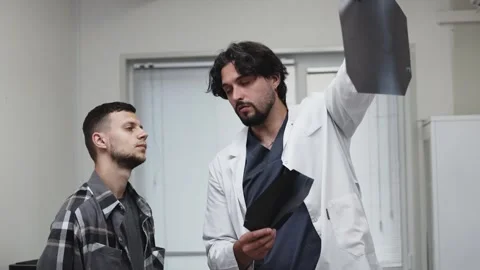 Doctor in white coat explaining knee X-ray to patient in gray plaid shirt, both Stock Footage 339980021