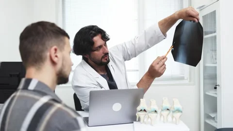 Doctor in white coat explaining knee X-ray to male patient, pointing with pen at Stock Footage 339980428