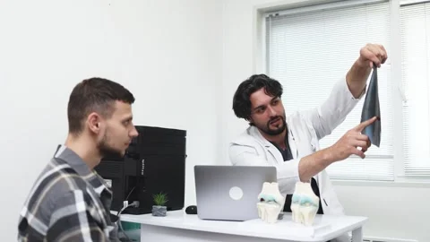 Doctor in white coat explaining knee X-ray to male patient seated at desk with Stock Footage 339980593