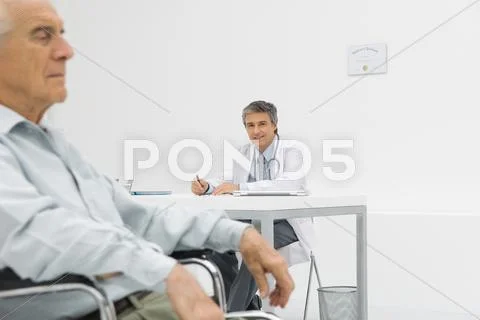 Doctor Writing At Desk In Office, Smiling At Camera, Patient Sitting In