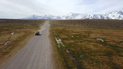 Dog runs down a remote dirt road with truck, dust cloud, and mountains behind 스톡 동영상 89857296