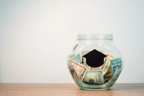 Dollar banknotes in the glass jar on the table, black student`s hat on it Stock Photos