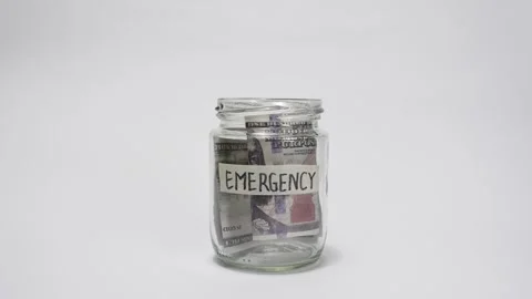Dollar bills falling into a glass jar labeled Emergency on a white background Stock Footage 316983825
