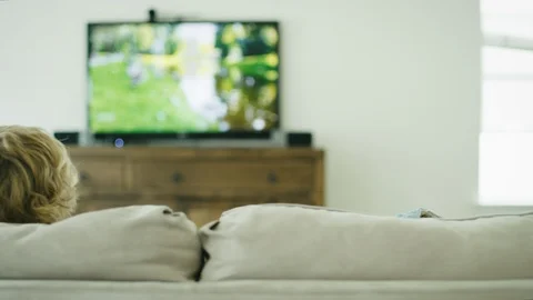 Dolly behind blonde boy on couch watching video of himself on television 스톡 동영상 127549978