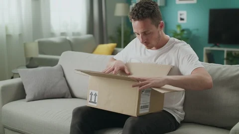 Dolly out of young man opens a cardboard parcel on the couch in his living room Stockbeeldmateriaal 108549276