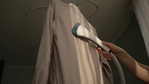 Domestic worker using garment steamer for steaming of wrinkle-free beige shirt Stock Footage 305509838