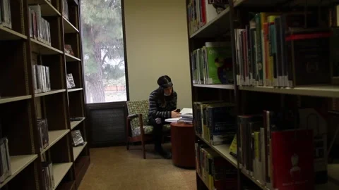 Down the aisle looking at teen in library with books, medical mask pulled down Stock Footage 134672301