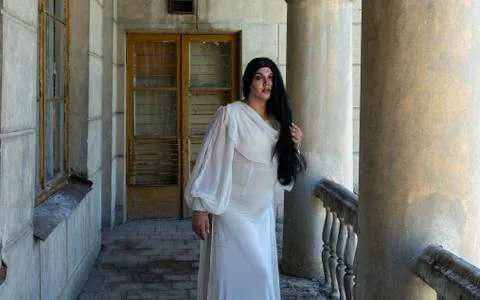 Drag queen posing close to old building in white dress. A wig with Black long 스톡 사진