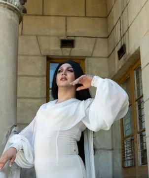 Drag queen posing close to old building in white dress. A wig with Black long Stock Photos