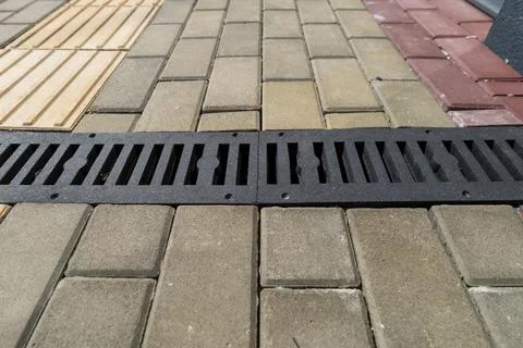 A drain cover sits between paver stones on a sidewalk in a city. The scene .. Stock Photos