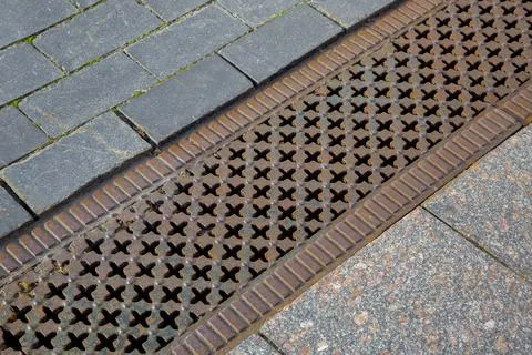 A drainage ditch covered with an iron patterned lattice on a sidewalk. Stock Photos