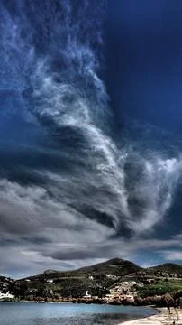 Dramatic cirrus cloud formation over coastal bay and hillside village in Kea Stock Photos