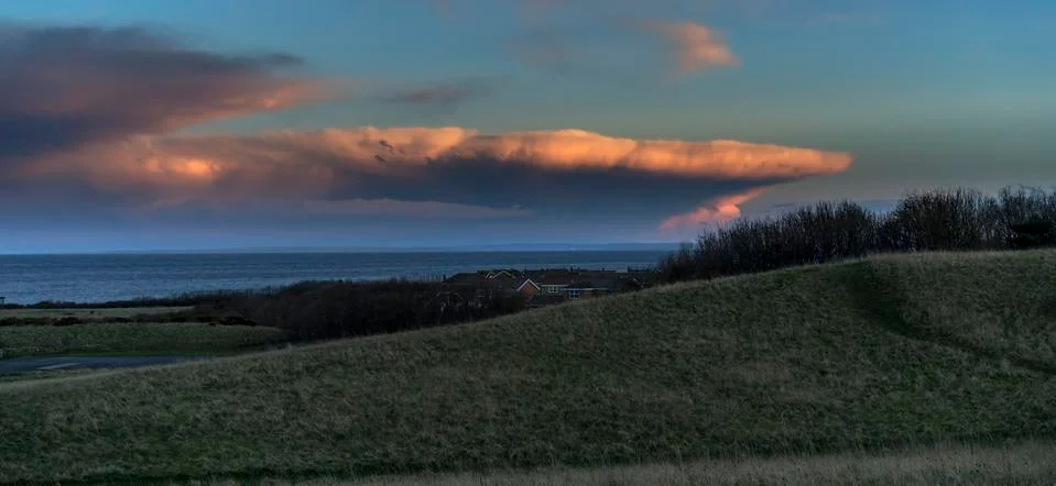 Dramatic cloud formation glowing pink at sunset over the ocean with green fields Stock Photos