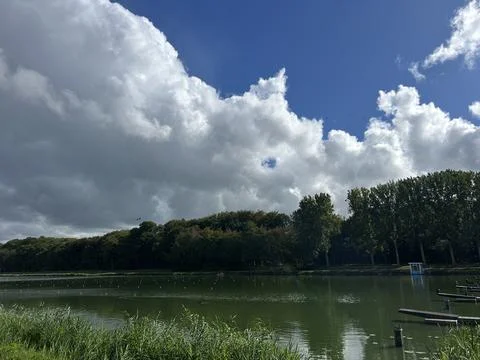Dramatic cloudscape over tree-lined lake with red buoy markers, reeds on grassy Stock-Fotos