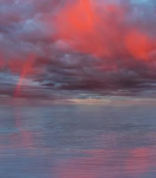 Dramatic cloudy sky with rainbow and red rain curtains reflecting on sea Stock Photos