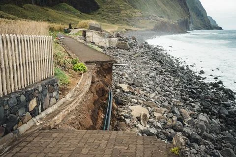 Dramatic coastal erosion scene with a damaged path, fence, and rocky beach. A Stock Photos