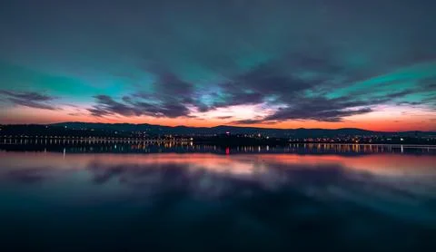 Dramatic colorful sunset reflection over calm lake with city lights, scenic Photos