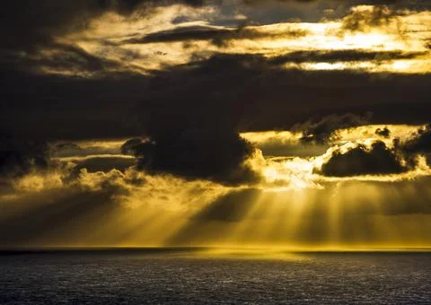 Dramatic golden sunbeams break through dark storm clouds over the ocean 写真素材