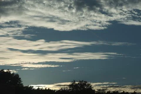 Dramatic layered clouds fill a blue evening sky above silhouetted trees and.. Stock Photos