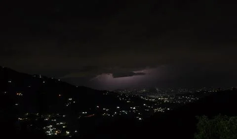 Dramatic lightning storm over a mountainous town at night with glowing city. Stock Photos
