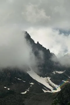 Dramatic mountain peak obscured by clouds with snow and rainbow Stock Photos