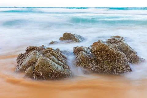 Dramatic moving wave around big rocks on clear water beach. Stock Photos