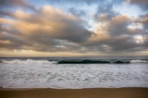 Dramatic ocean waves crash on a sandy beach under a cloudy sunset sky Photos