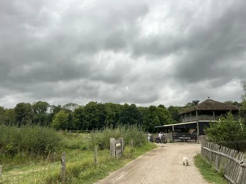 Dramatic overcast sky over rural park path with wooden lodge, cyclists near Stock Photos