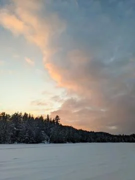 Dramatic Pastel Sunset Clouds Over Frozen Snow Covered Lake and Winter Forest Stock Photos