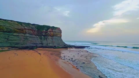 Dramatic rocky cliff bordering wet sandy beach at sunset as ocean waves crash Stock Footage 345483709