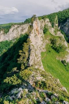 Dramatic rocky cliff surrounded by lush forest and green hills under a brig.. Stock Photos