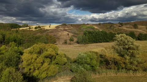 Dramatic storm clouds hang over rolling golden hills and dense green forests in Видео 347160513